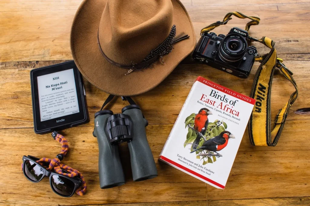 Flat lay of safari travel essentials including binoculars, field guide book, hat, and camera.